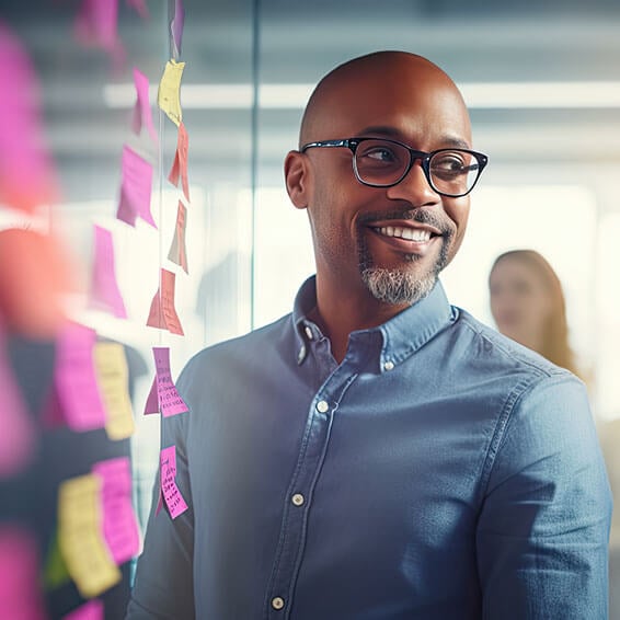 A smiling man wearing glasses stands in front of a colorful wall covered in sticky notes