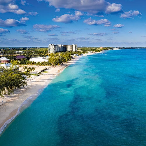 Aerial view of Seven Mile Beach in Grand Cayman, showing the white sandy beach and a resort in the distance under a clear blue sky