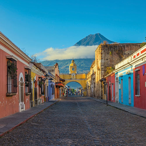 A vibrant street in Antigua, Guatemala, lined with colorful colonial buildings, leading to the iconic Arco de Santa Catalina arch and the majestic Volcano de Agua in the background