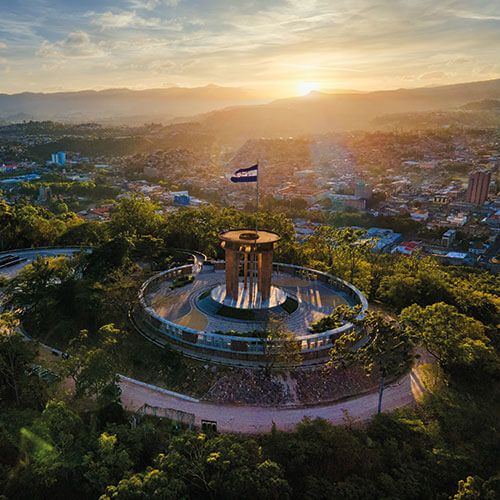 Aerial view of the Monumento a la Madre in Tegucigalpa, Honduras, with a Honduran flag waving at sunset and the city skyline