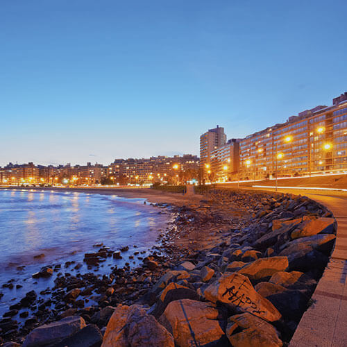 An aerial shot of the Rambla de Montevideo at dusk with illuminated coastline, promenade, and beach