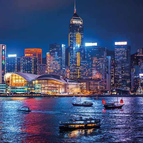 A panoramic view of Hong Kong skyline at night with illuminated skyscrapers and boats in the harbor