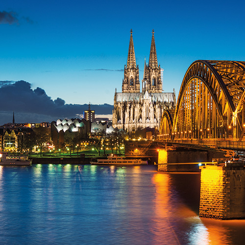 A scenic view of the Cologne Cathedral and Hohenzollern Bridge illuminated at night, overlooking the Rhine River