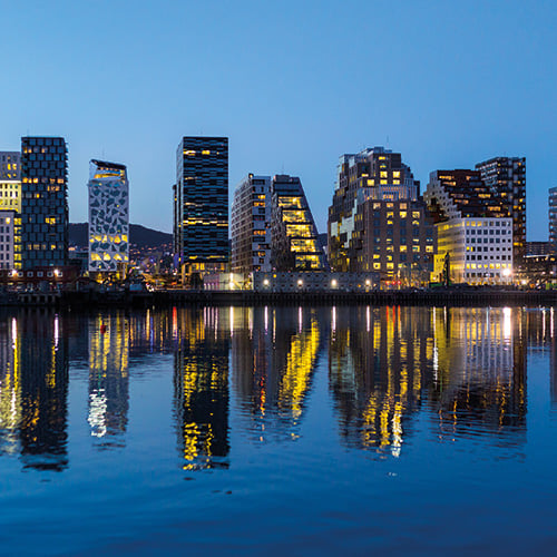 A beautiful image of the Oslo waterfront at night, featuring a row of modern skyscrapers illuminated against a dark sky