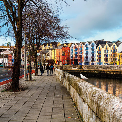 A scenic view of colorful buildings lining the River Lee in Cork, Ireland, with pedestrians walking along the waterfront