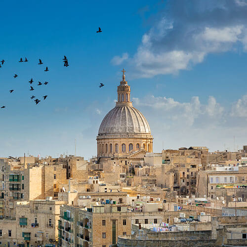 A panoramic view of Valletta, Malta, with St. John's Co-Cathedral dome and St. Paul's Anglican Pro-Cathedral spire, against a blue sky with birds flying overhead