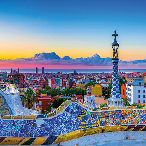 A panoramic view of Barcelona, Spain, from Park Güell, featuring colorful mosaic benches, iconic architecture, and a beautiful sunrise over the city