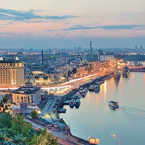 A panoramic view of Kyiv, Ukraine, at twilight, featuring the Dnieper River, the illuminated city skyline, and the iconic Hotel Rus