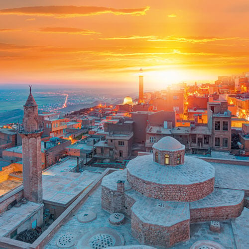 A panoramic view of Mardin, Turkey, at sunset, showcasing the historic city's stone buildings, minarets, and the surrounding landscape