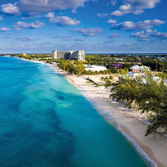 An aerial view of Seven Mile Beach in Grand Cayman, showing a pristine white sandy beach and a resort in the distance under a clear blue sky