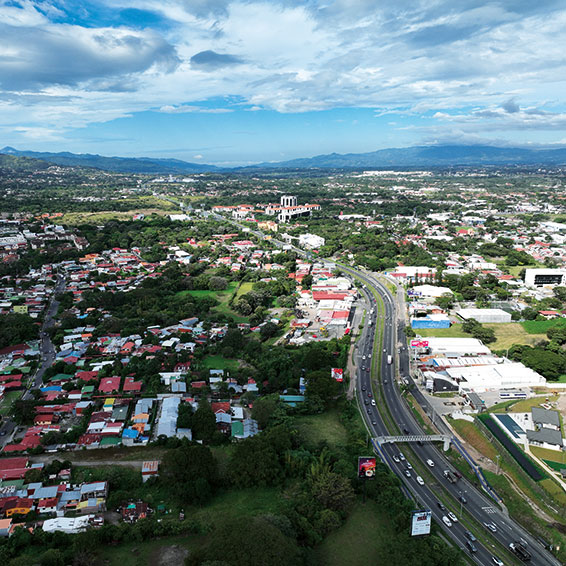 An aerial view of Santa Ana, Costa Rica, showing a suburban area with a highway, green spaces, and mountains in the distance