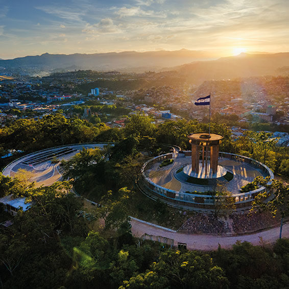 An aerial view of the Monumento a la Madre in Tegucigalpa, Honduras, with a Honduran flag waving at sunset and the city skyline in the background