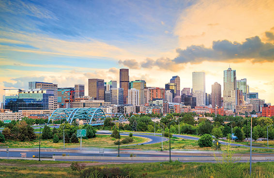 A panoramic view of the Denver skyline at sunset, with a bridge and road leading to the city