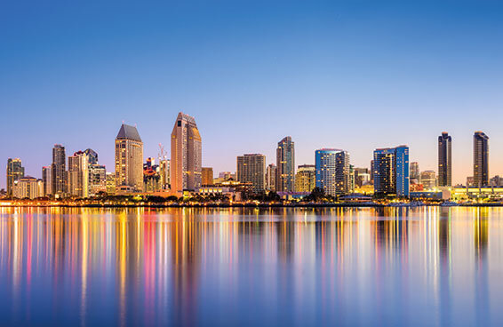 A panoramic view of the San Diego skyline at dusk, with the city reflected in the calm water