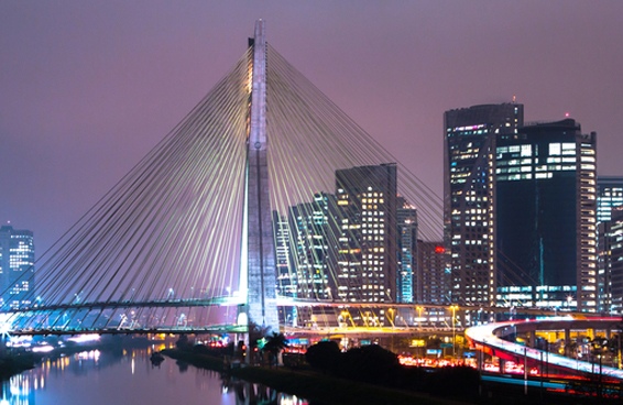 A scenic view of the Octavio Frias de Oliveira Bridge illuminated at night, with the São Paulo skyline in the background