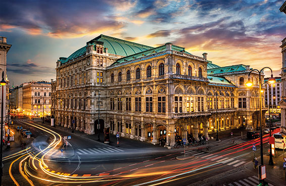 An aerial view of the Vienna State Opera at dusk, with traffic lights creating streaks in the foreground