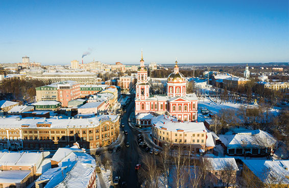 An aerial view of Kirov, Russia, featuring a red and white Orthodox church amid snow-covered streets and buildings under a clear blue sky