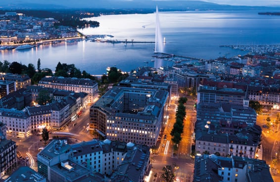 An aerial view of Geneva at dusk, with Lake Geneva, the Jet d'Eau fountain, and the city's illuminated streets