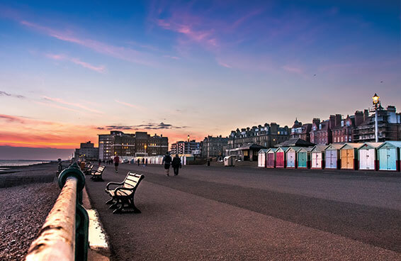 A view of the seafront in Brighton, England, at sunset, with colorful beach huts and people walking along the promenade