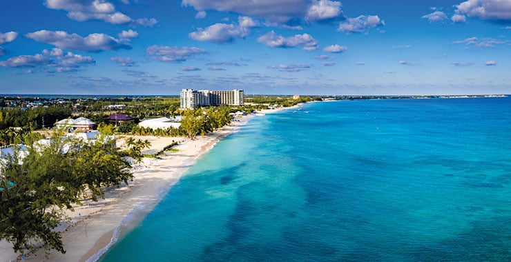 An aerial view of Seven Mile Beach in Grand Cayman, showing a pristine white sandy beach and a resort in the distance under a clear blue sky