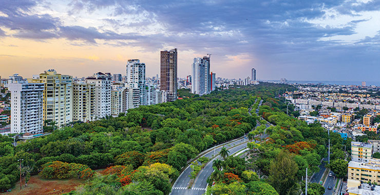 An aerial view of Santo Domingo, Dominican Republic, showing a cityscape with tall buildings and a green park with a road winding through it under a dramatic cloudy sky