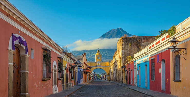 A shot of the vibrant street in Antigua, Guatemala, lined with colorful colonial buildings, leading to the iconic Arco de Santa Catalina arch and the majestic Volcano de Agua in the background