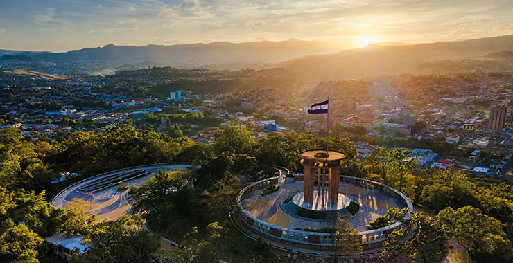 An aerial view of the Monumento a la Madre in Tegucigalpa, Honduras, with a Honduran flag waving at sunset and the city skyline in the background