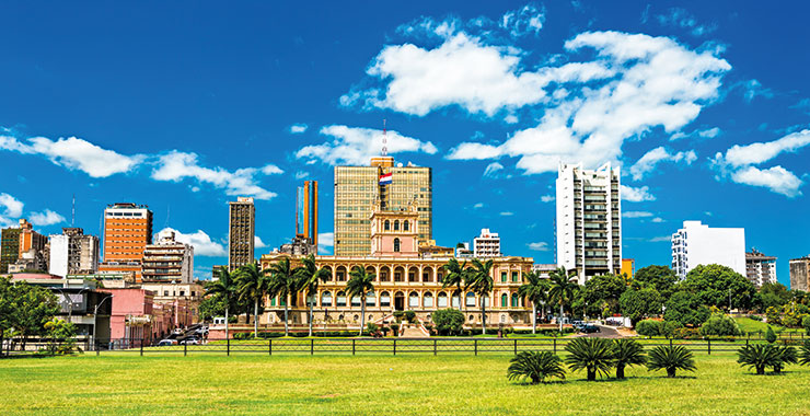 A panoramic view of the Palacio de López in Asunción, Paraguay, with a Paraguayan flag flying and lush green lawns in the foreground