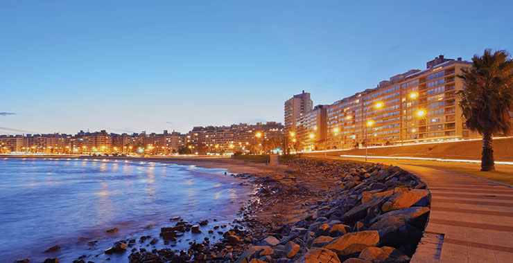 An aerial shot of the Rambla de Montevideo at dusk, with illuminated coastline, promenade, and beach