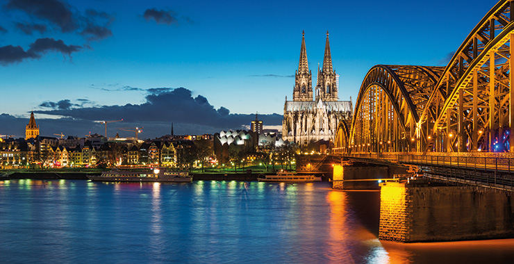 A scenic view of the Cologne Cathedral and Hohenzollern Bridge illuminated at night, overlooking the Rhine River