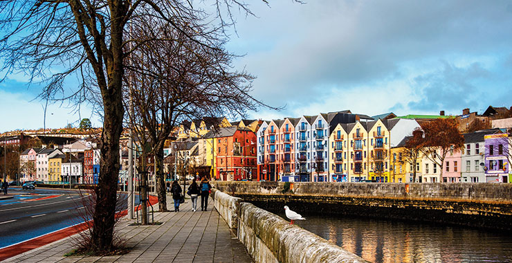 A scenic view of colorful buildings lining the River Lee in Cork, Ireland, with pedestrians walking along the waterfront