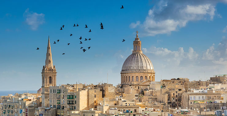 A panoramic view of Valletta, Malta, with St. John's Co-Cathedral dome and St. Paul's Anglican Pro-Cathedral spire, against a blue sky with birds flying overhead