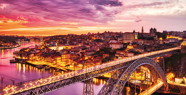 A panoramic view of Porto, Portugal, at sunset, featuring the Dom Luís I Bridge illuminated over the Douro River