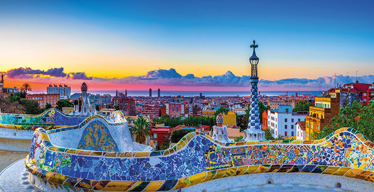 A panoramic view of Barcelona, Spain, from Park Güell, featuring colorful mosaic benches, iconic architecture, and a beautiful sunrise over the city