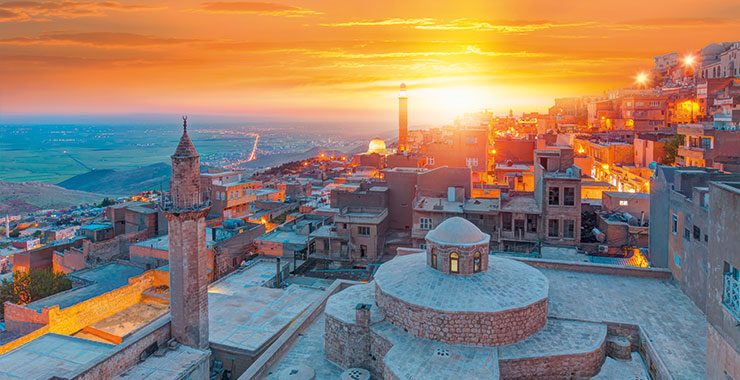 A panoramic view of Mardin, Turkey, at sunset, showcasing the historic city's stone buildings, minarets, and the surrounding landscape