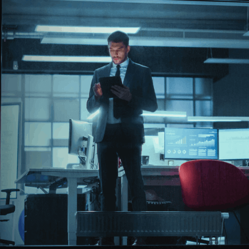 Businessman in a dimly lit office reviewing a tablet near computer screens